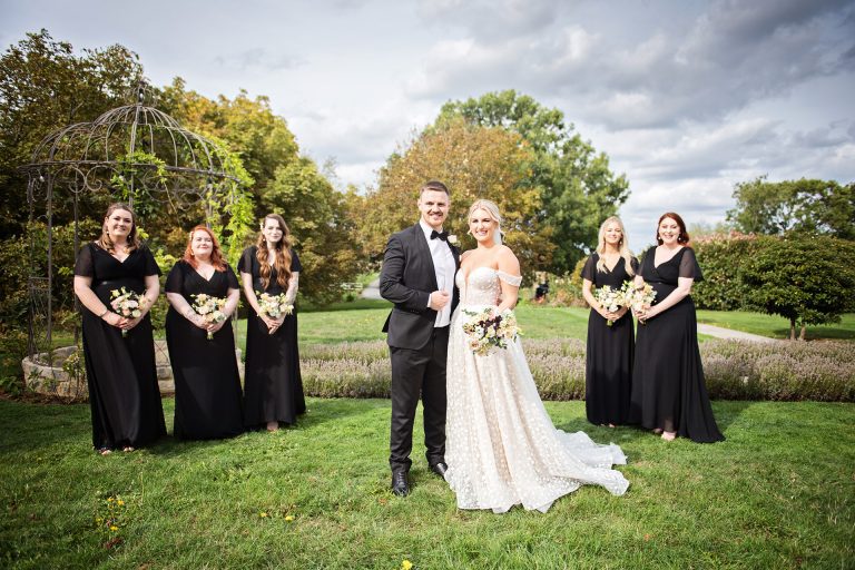 Bridesmaids in their black dresses, and the bride and groom looking forward at the camera. Autumnal trees behind them. Photo by Blooming Photography taken at Blackwell Grange, Warwickshire wedding.