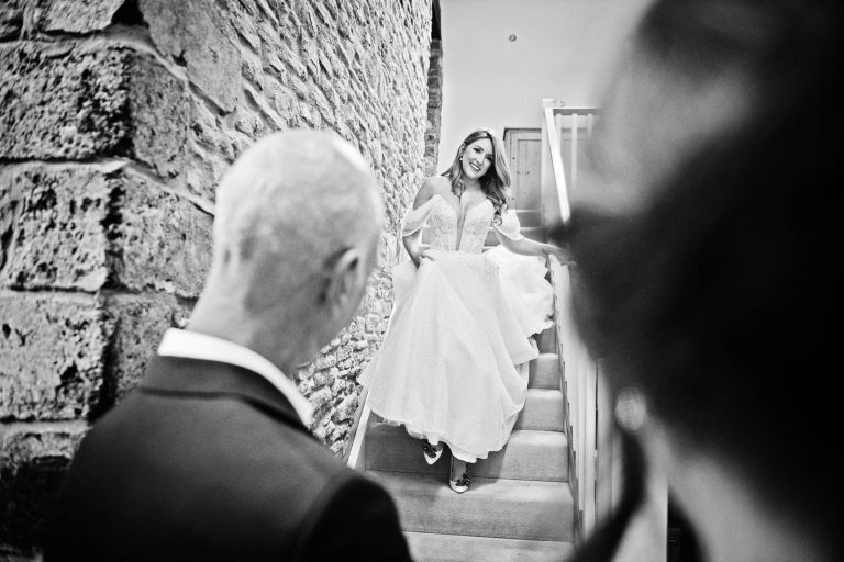 Candid black and white photo of bride walking down the stairs to be greeted by her dad. Cardiff wedding photographer, Blooming Photography