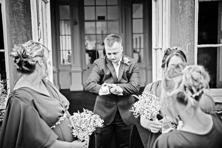 Black and white candid photo of a groomsman looking at his watch with bridesmaids either side of him. Photographed by Blooming Photography at Margam Country Park.