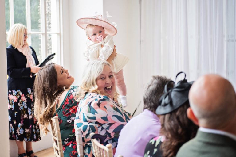 Candid pic of baby wearing a wedding fascinator being held up in the air taken by Blooming Photography at a wedding at Margam Country Park.