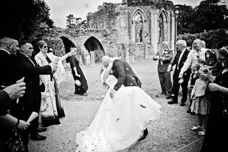 Candid black & white photo of a bride and groom doing a wedding dip whilst having wedding confetti thrown at them. Photo taken at a wedding at Margam Country Park by Blooming Photography.