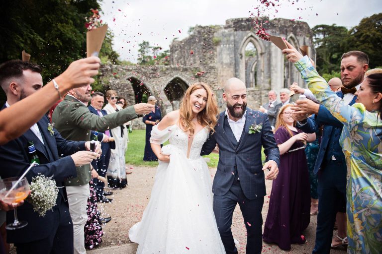 Candid colour photo of a bride and groom smiling whilst having wedding confetti thrown at them. Photo taken at a wedding at Margam Country Park by Blooming Photography.
