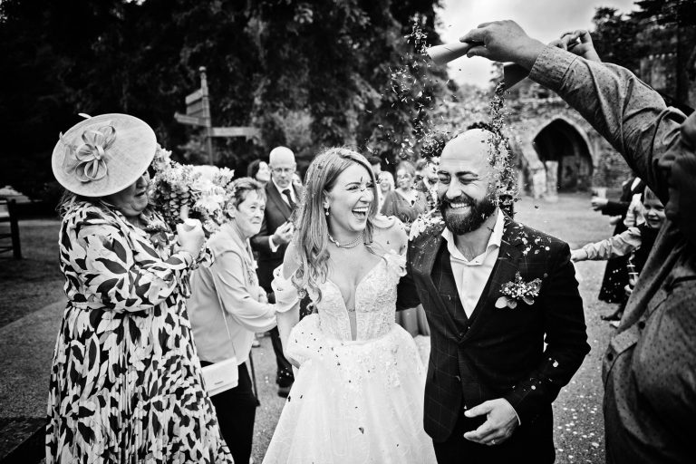 Natural black & white photo of a bride and groom smiling whilst having wedding confetti thrown at them. Photo taken at a wedding at Margam Country Park by Blooming Photography.