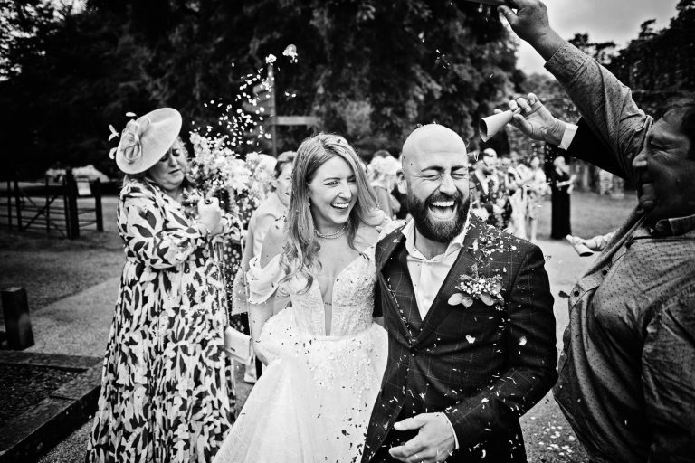 Candid black & white photo of a bride and groom smiling whilst having wedding confetti thrown at them. Photo taken at a wedding at Margam Country Park by Blooming Photography.