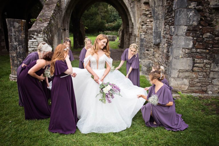 Bridesmaids gather the brides wedding dress with the ruins behind them. Photo taken at Margam Country Park by Blooming Photography.