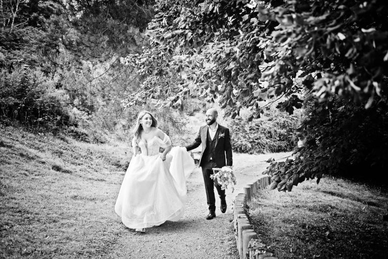 Natural black & white wedding photo of bride and groom walking up smiling whilst the groom is holding the brides dress up. Photo taken at Margam Country Park by Blooming Photography.