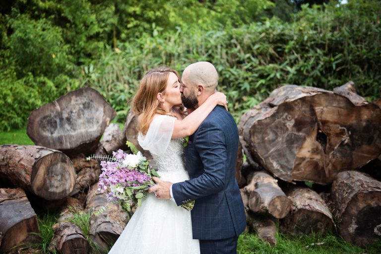 Photo of bride and groom holding each other closely,, kissing with logs behind them at Margam Country Park behind them - photo by Blooming Photography.
