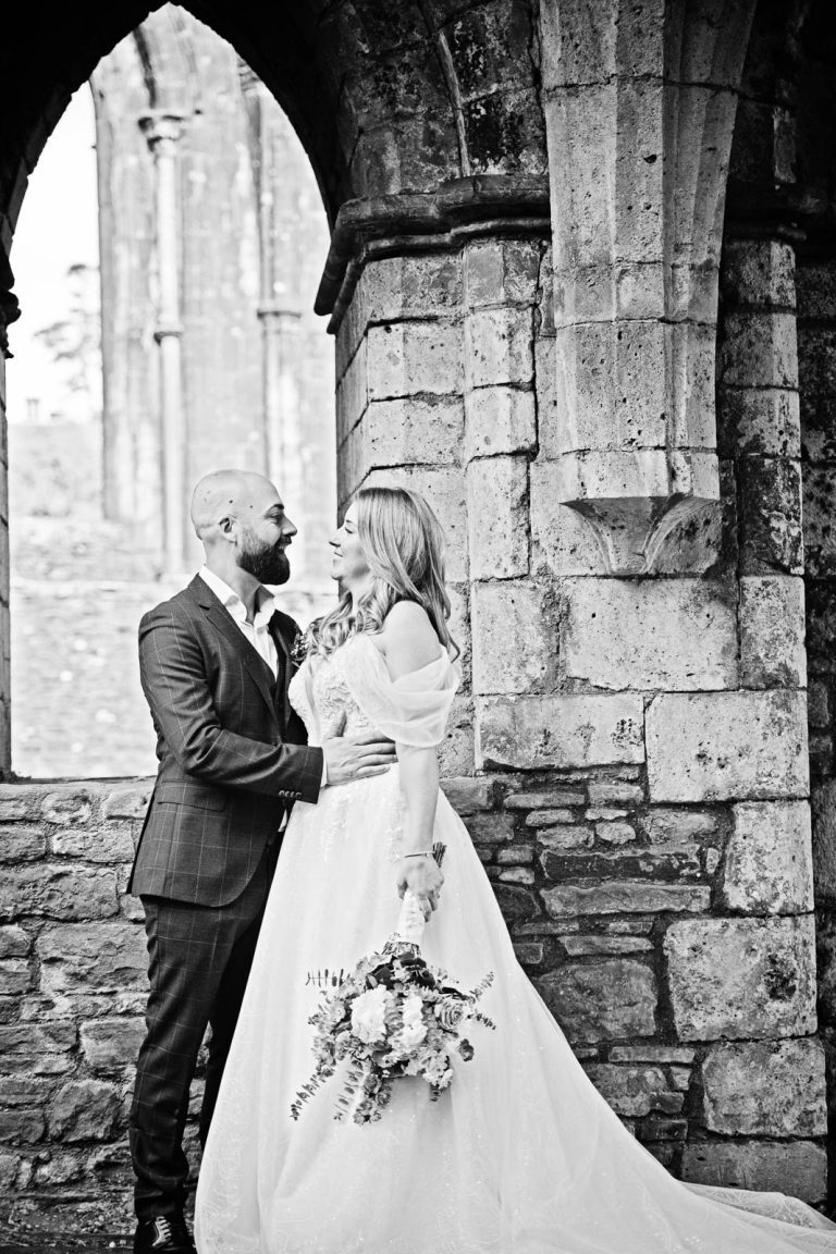 Candid B& W wedding photo of bride and groom holding each other closely, smiling with the ruins at Margam Country Park behind them - photo by Blooming Photography.
