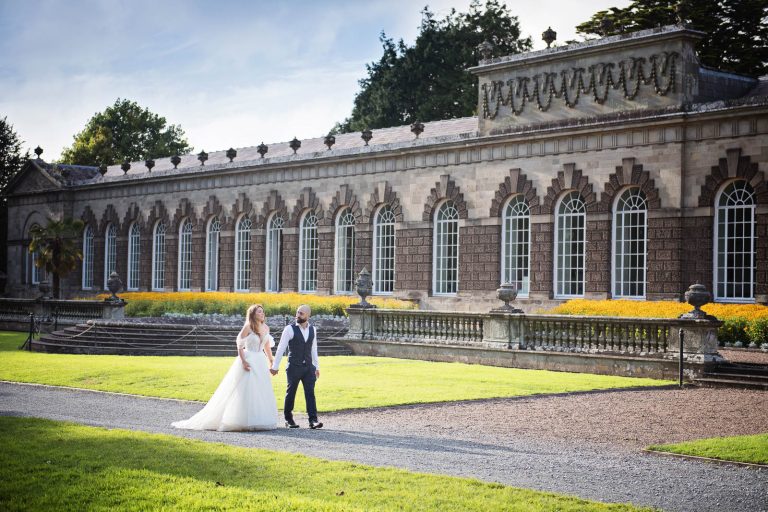 Elegant wedding photograph of bride and groom holding hands and walking down a path with Margam Country Park Hall behind them, photo by Blooming Photography.