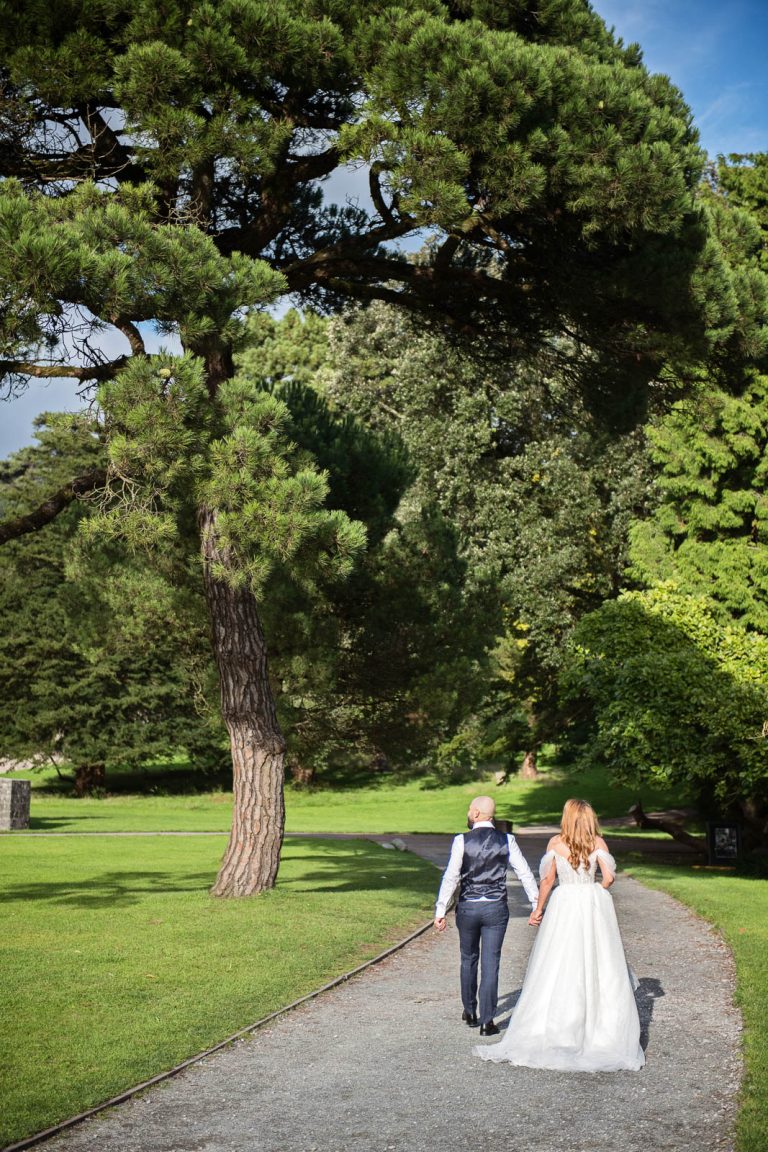 Relaxed wedding photography of bride and groom holding hands and walking away down a path at Margam Country Park photo by Blooming Photography.