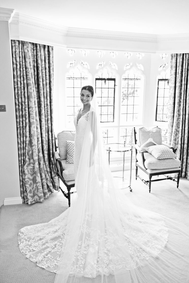Bride looking over her shoulder in her wedding dress. Standing in front of a historic window. Photography by Blooming Photography at Danesfield House Hotel and Spa.