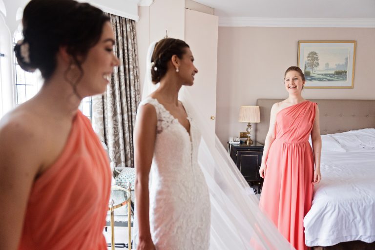 Bridesmaid and bride looking toward another bridesmaid both smiling. Candid. Photography by Blooming Photography at Danesfield House Hotel and Spa.