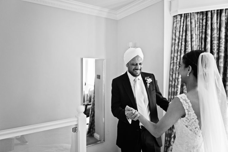 Father of the bride seeing his daughter for the first time. He is holding the brides hands and smiling. Photography by Blooming Photography at Danesfield House Hotel and Spa.