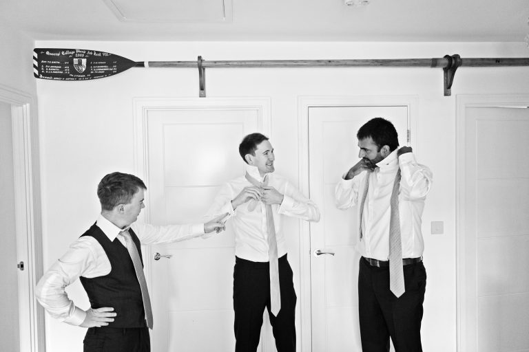 Candid black and white photo of three groomsmen tying their ties, with a Queens College Rowing oar above them. Photography by Blooming Photography.