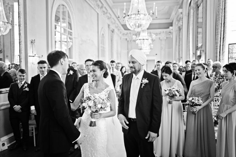Photo of the bride meeting the groom for the first time after she has walked down the wedding isle at Danesfield House. Father of the bride and all guests look emotional. Black and white photograph. Photography by Blooming Photography.
