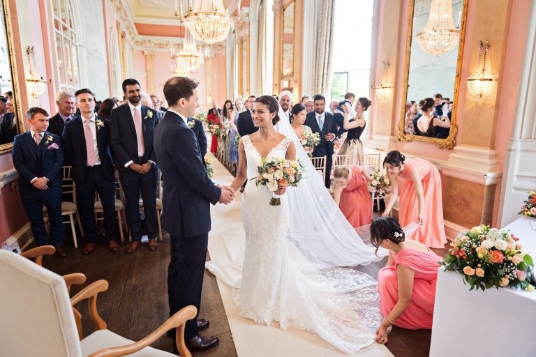 Bride and groom hold hands as they meet at the wedding aisle. Bridesmaids are spending time to correctly lay out the dress and veil. Photography by Blooming Photography at Danesfield House Hotel and Spa.