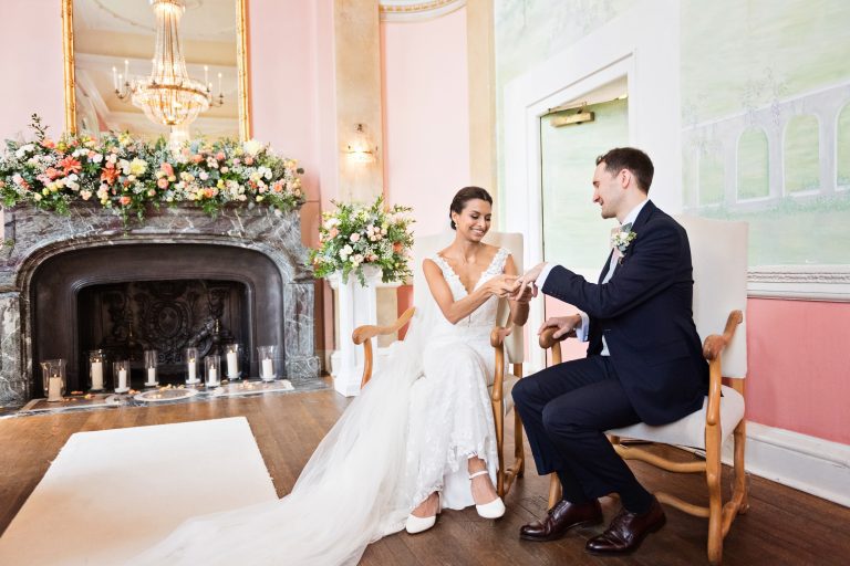 Bride and groom exchange rings whilst sitting down in stately chairs. A gorgeous fireplace with flowers and candles and a mirror reflecting a chandelier adds to the quality of the image. Photography by Blooming Photography at Danesfield House Hotel and Spa.