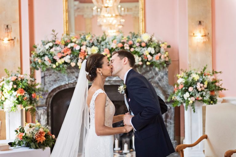 A bride and groom exchange a kiss after being announced "Husband and wife". A sea of stunning wedding flowers are behind them. Photography by Blooming Photography at Danesfield House Hotel and Spa.