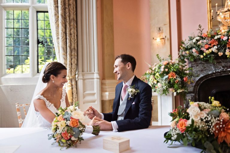A bride and groom hold hands and smile and laugh at each other after signing the marriage certificate. Photography by Blooming Photography at Danesfield House Hotel and Spa.