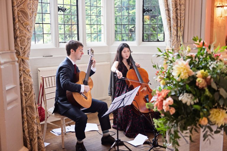 An acoustic guitarist and cello player playing emphatic music. A bouquet of flowers in the foreground. Photography by Blooming Photography at Danesfield House Hotel and Spa.