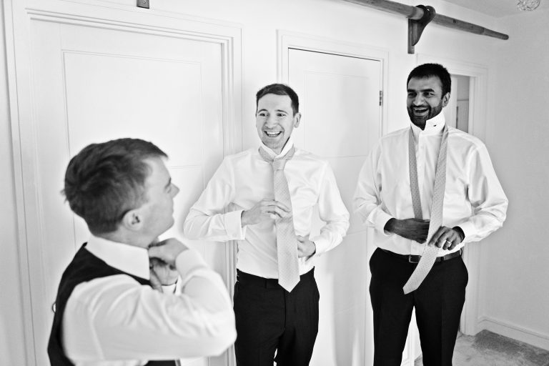 Candid black and white photo of three groomsmen tying their ties smiling. Photography by Blooming Photography.