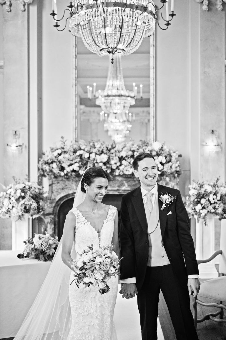 An elegant portrait image of a bride and groom holding hands and smiling with a row of chandeliers reflective in the mirror. Black and white image. Photography by Blooming Photography at Danesfield House Hotel and Spa.