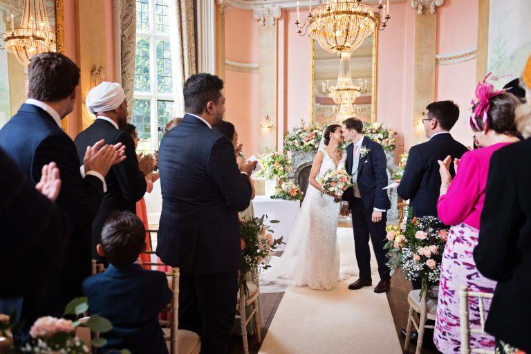 Wedding guests applaud the bride and groom who seal their marriage with a kiss in the grand hall at Danesfield House Hotel. Photography by Blooming Photography