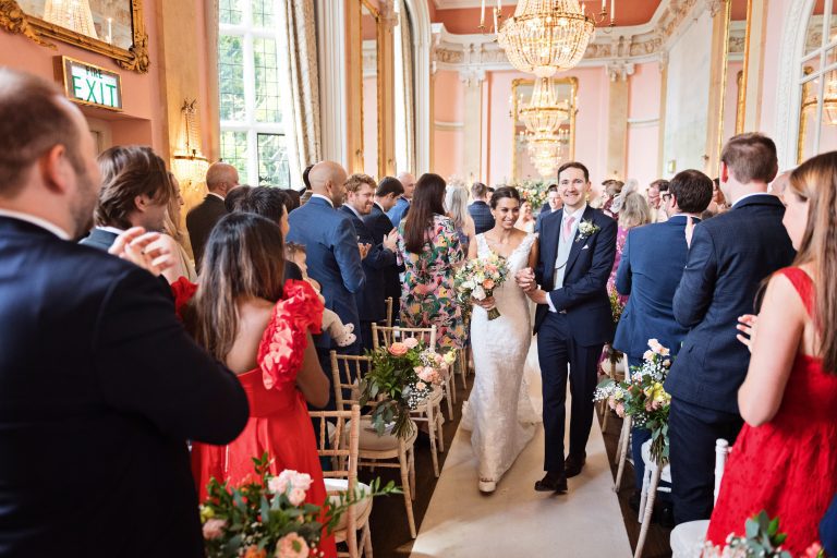 A bride and groom walk down the wedding isle together at Danesfield House Hotel and Spa with their wedding guests clapping. Elegant image. Photography by Blooming Photography.