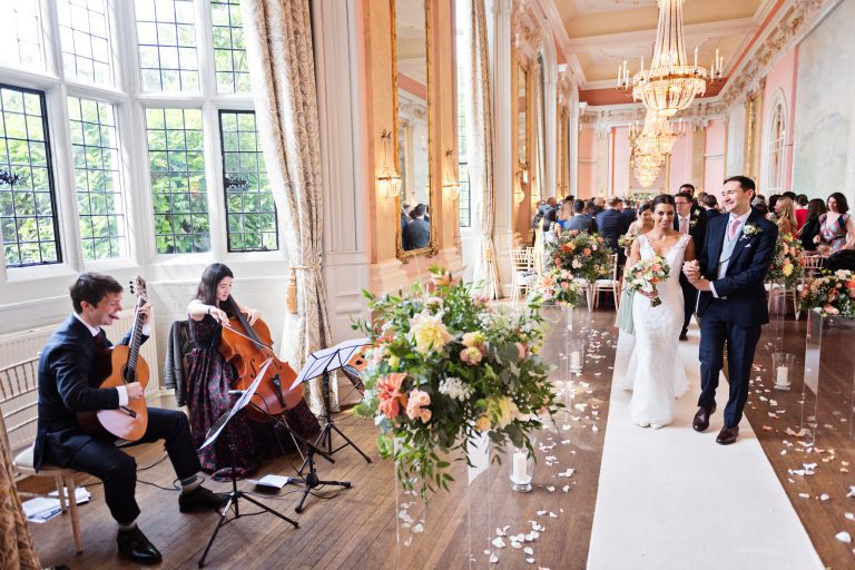 A bride and groom walk down the wedding isle together but pause at seeing the musicians play a beautiful classical piece of music at Danesfield House Hotel and Spa. Elegant image. Photography by Blooming Photography.