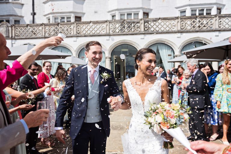 Photograph of the bride and groom (both smiling), walking down a confetti isle made by family and friends at Danesfield House Hotel and Spa. Photography by Blooming Photography.