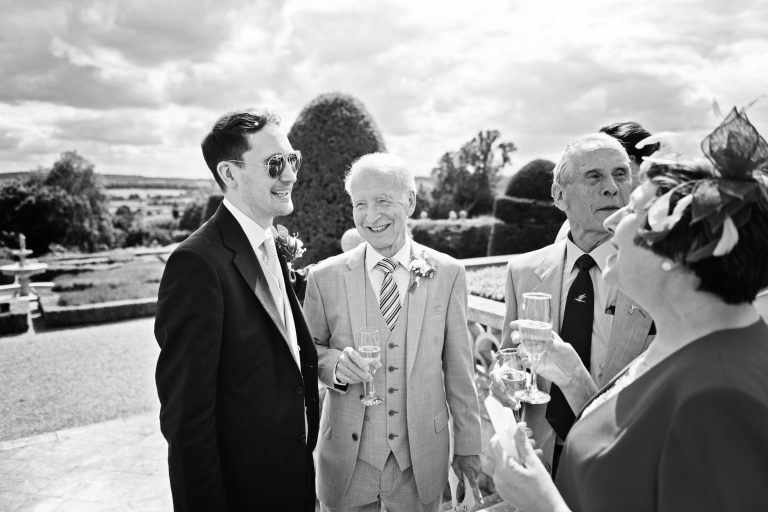 The groom chats to his parents informally, with the mother telling stories whilst the father is smiling in the gardens of Danesfield House Hotel and Spa. Black and white image. Photography by Ben Roberts, Blooming Photography.