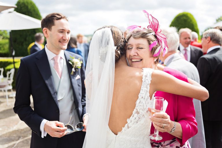 The mother of the groom hugs the bride with the groom onlooking. All smiling. Colourful photograph. Photographed at Danesfield House Hotel and Spa. Photography by Ben Roberts Blooming Photography.