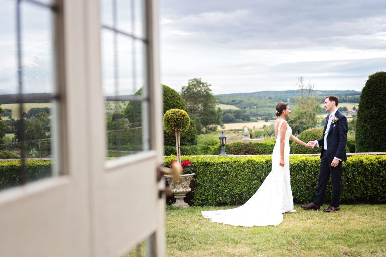 Bride and groom hold hands and have a moment together. Photographed candidly and composed as if looking through a window/door. Photographed at Danesfield House Hotel and Spa. Photography by Ben Roberts Blooming Photography.