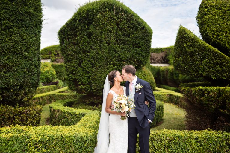 Bride and groom kiss with the magnificent topiary of Danesfield House behind them. Photography by Ben Roberts Blooming Photography.