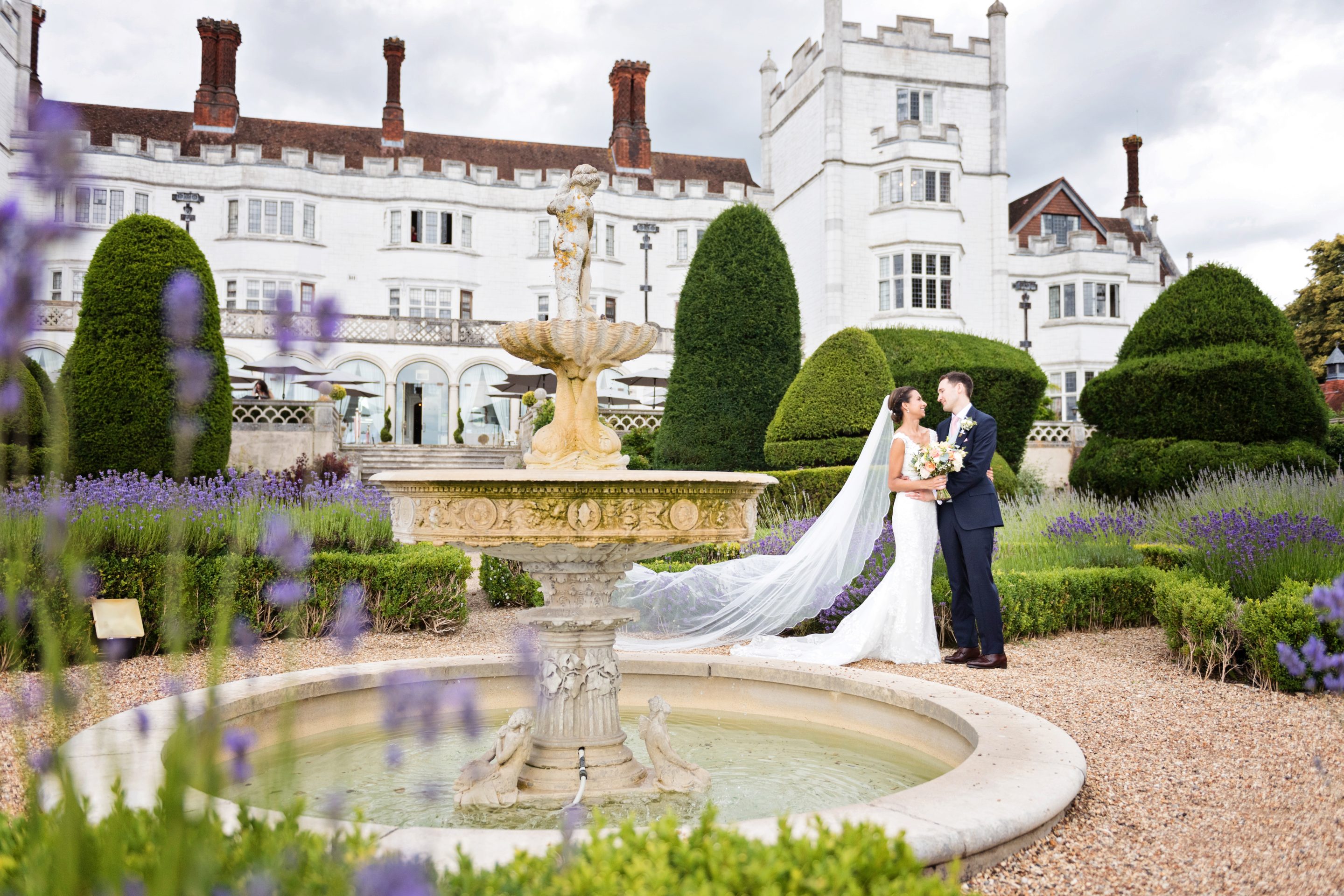 Bride and groom in the grounds at Danesfield House Hotel and Spa. Buildings behind them. Bride and groom stand behind the water fountain.