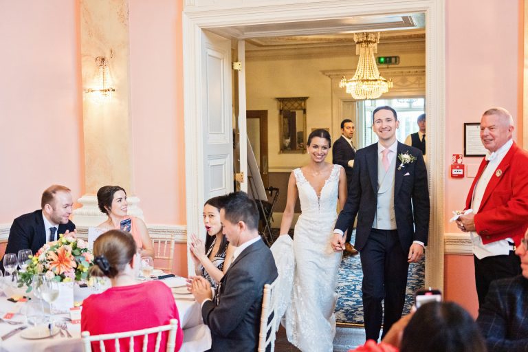 Bride and groom enter the wedding breakfast room at Danesfield House Hotel and Spa smiling with the friends and family smiling and clapping and taking photographs with their mobile phones. Photograph by Ben Roberts, Blooming Photography.