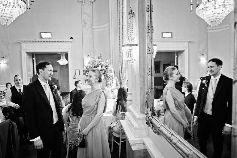 Groom chats with bride (times two) as image is doubled by using a mirror. Black and white. Chandelier in the ceiling. Danesfield House Ballroom. Photograph by Ben Roberts, Blooming Photography.