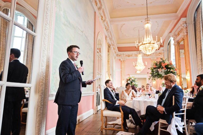 Bestman at wedding does his bestman speech. Guests (including the bride and groom) are laughing. Danesfield House Ballroom. Photograph by Ben Roberts, Blooming Photography.
