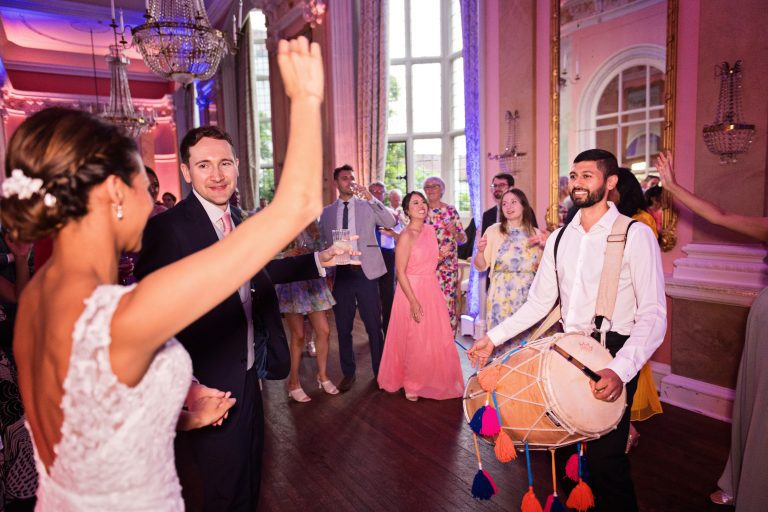 Bride and groom greet the Indian drummers. Photographed in the elegant Danesfield House Hotel and Spa Ballroom. Photograph by Ben Roberts, Blooming Photography.