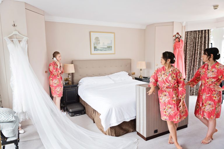 Bridesmaids getting the call to say you need to get ready by the hotel. Dress is laid out elegantly in between them. Photography by Blooming Photography at Danesfield House Hotel and Spa.