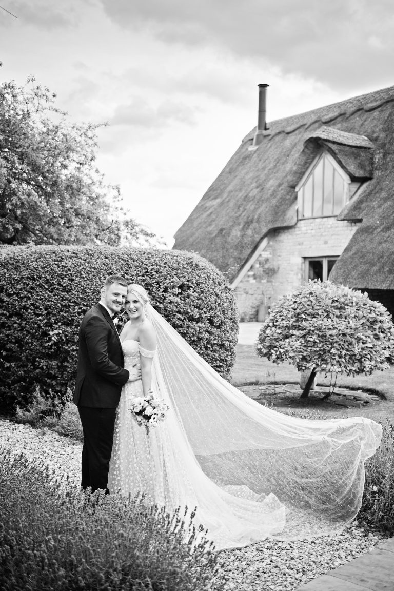 B&w image of bride and groom smiling towards the camera. A thatched building behind them. Candid photo by Blooming Photography taken at Blackwell Grange, Warwickshire wedding.