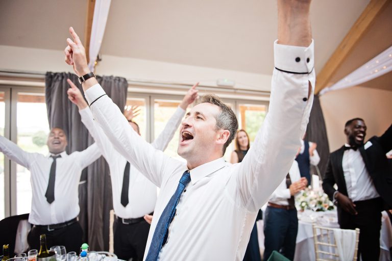 Candid photo by Blooming Photography. Taken at Blackwell Grange, Warwickshire. Wedding guests cheer and sing with their hands in the air. Party!