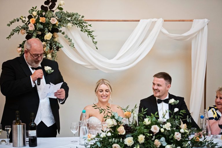 Father of the bride delivering his speech with the bride and groom smiling next to him. Candid photo by Blooming Photography. Taken at Blackwell Grange, Warwickshire.