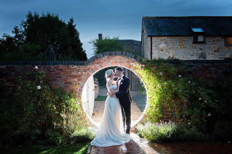 bride and groom kiss together whilst being lit up by a light through a circular brick doorway. Elegant photo by Blooming Photography. Taken at Blackwell Grange, Warwickshire.
