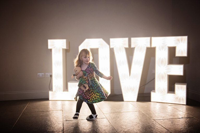 Love letter lights with a child holding a baby doll in front of the sign. Candid photo by Blooming Photography. Taken at Blackwell Grange, Warwickshire.