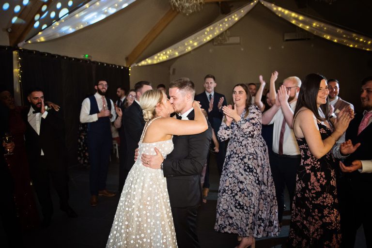 Bride and groom kiss with their guests clapping after they have finished their first dance. Candid photo by Blooming Photography. Taken at Blackwell Grange, Warwickshire.