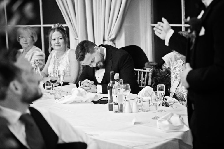 Groom hangs his head (but smiling) during the best man speech. Black and white photograph by Blooming Photography taken at Eastington Park at Christmas Wedding.