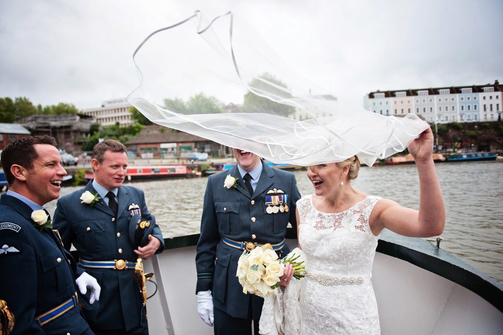 Candid photo of Bride and groom (dressed in military uniform) on a boat in Bristol after their wedding smiling/laughing with two of their guests as the wind blows the veil in the air in front of them. Photo by Blooming Photography