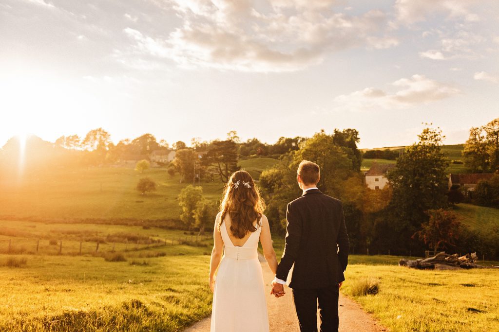 Bride and groom with their backs to the camera, holding hands looking back on Kingscote Barn, Photo by Gloucestershire wedding photographer Blooming Photography.