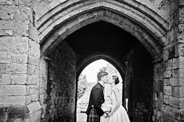 Candid wedding photo of bride and groom kissing under medieval arch Winchester, photo by Blooming Photography.